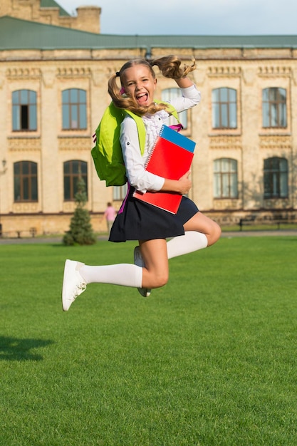 Ragazza carina salta con notebook felice ragazza in età prescolare con libro nel cortile della scuola torna a scuola bambino laborioso portare zaino concetto di istruzione vacanze primaverili vacanze estive