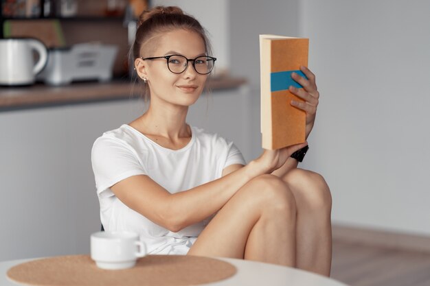 Ragazza carina sta bevendo caffè e leggendo un libro al tavolo della cucina a casa