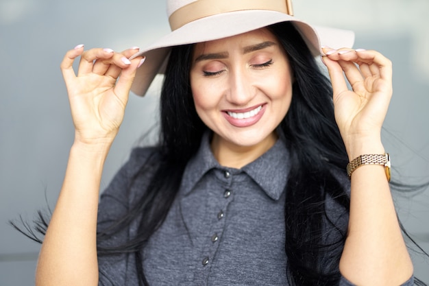 Ragazza castana attraente allegra del ritratto del primo piano con gli occhi chiusi che portano tenuta grigia alla moda del vestito e cappello alla moda. Focalizzazione morbida.