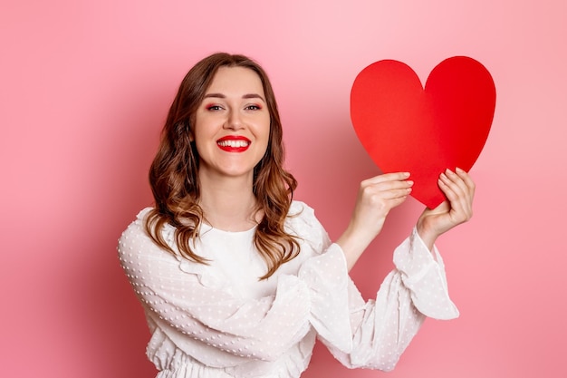 Ragazza castana caucasica in vestito bianco che tiene la carta di festa di San Valentino del grande cuore di carta