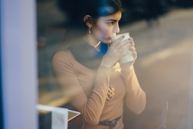 ragazza castana che si siede ad un caffe che beve una bevanda calda mentre guardando fuori la finestra