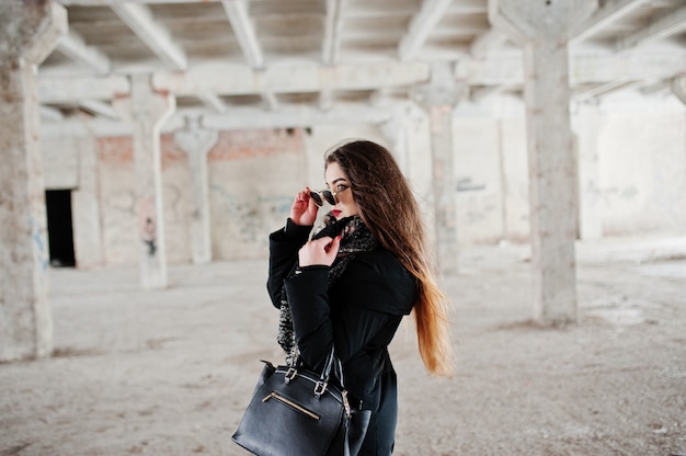 Ragazza casual alla moda castana in sciarpa e borsa contro il posto abbandonato della fabbrica.