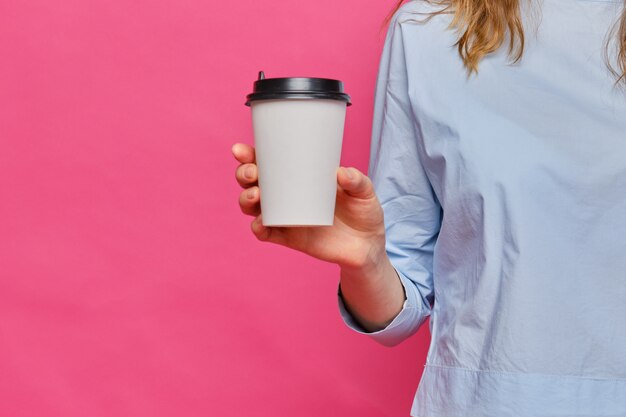 Ragazza caucasica alla moda in una maglietta blu-chiaro che tiene una tazza di carta per caffè su un fondo rosa.