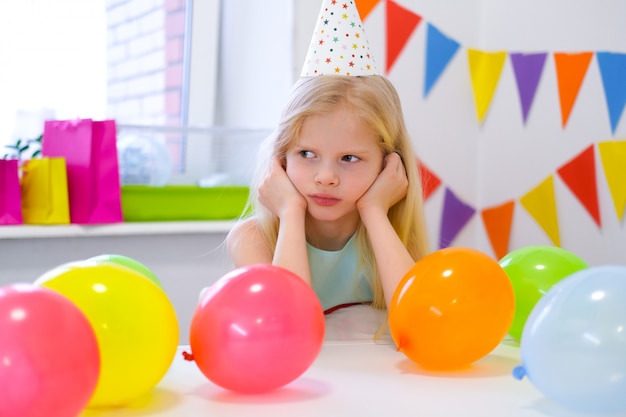 Ragazza caucasica bionda infelice con il fronte noioso vicino alla torta dell'arcobaleno di compleanno. Sfondo colorato festivo. Brutta festa di compleanno