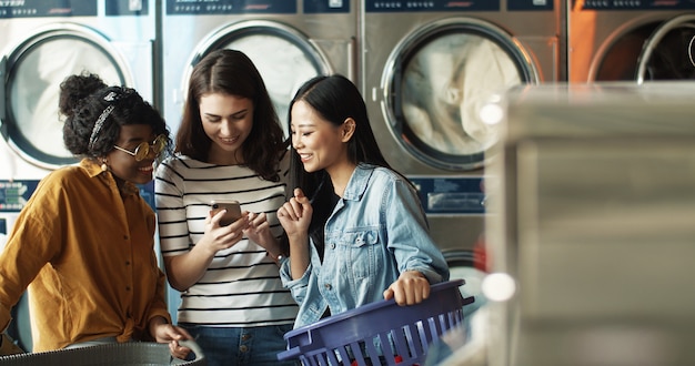 Ragazza caucasica che mostra le foto sullo smartphone agli amici femminili di razza mista mentre lavatrici lavorano e puliscono i vestiti. Donne multietniche che guardano video sul telefono nel servizio di lavanderia.