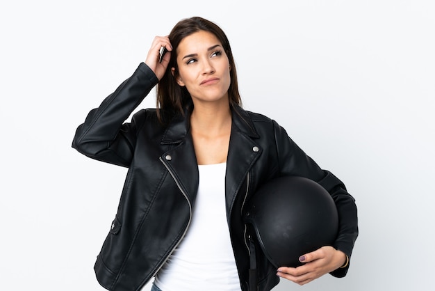 Ragazza caucasica che tiene un casco del motociclo su priorità bassa bianca che ha dubbi e con l'espressione confusa del fronte