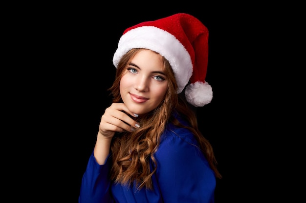 Ragazza caucasica con i capelli lunghi in abito blu che indossa il cappello di Babbo Natale isolato su sfondo nero