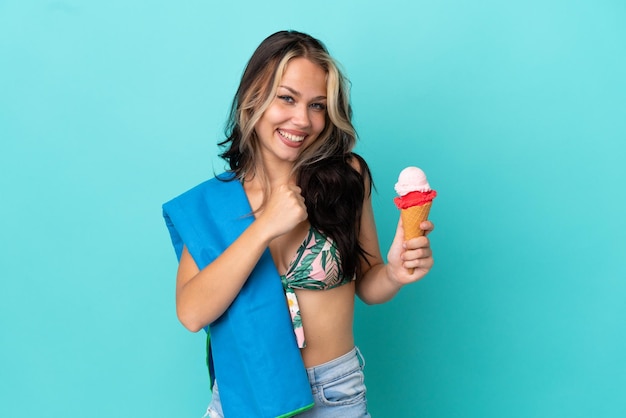 Ragazza caucasica dell'adolescente che tiene il gelato e l'asciugamano isolati su fondo blu che celebra una vittoria