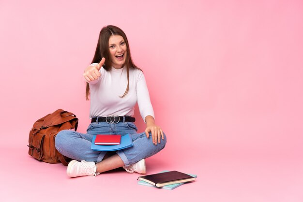 Ragazza caucasica dell'allievo dell'adolescente che si siede sul pavimento isolato sul colore rosa con i pollici in su perché è successo qualcosa di buono