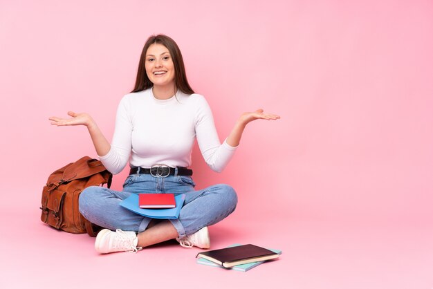 Ragazza caucasica dello studente dell'adolescente che si siede sul pavimento isolato sul rosa con espressione facciale colpita