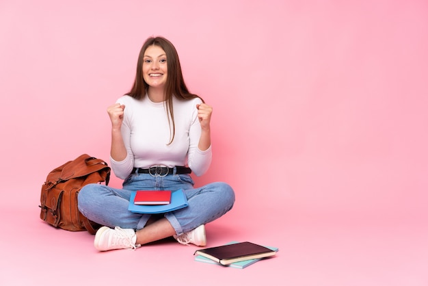 Ragazza caucasica dello studente dell'adolescente che si siede sul pavimento isolato sulla parete rosa che celebra una vittoria nella posizione del vincitore