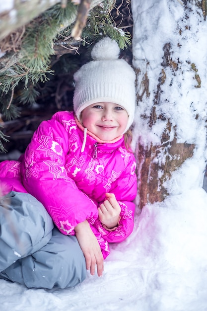 Ragazza caucasica di 5 anni che guarda la telecamera in una capanna da rami di conifere nella foresta invernale, giocando all'aperto in inverno