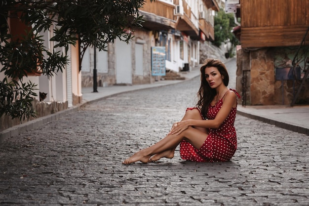 Ragazza caucasica in un vestito rosso che posa seduto su una pietra per lastricati su una bella strada europea