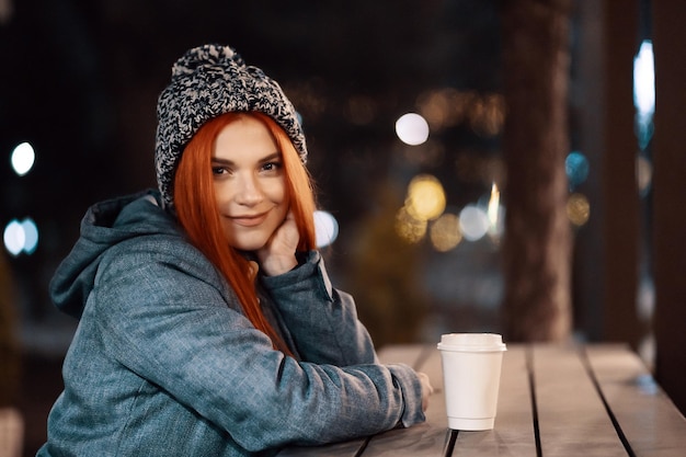 Ragazza che beve caffè caldo mentre è seduto al tavolo nel mercato serale della città decorato con luci natalizie. Sentirsi felici in una grande città. Trascorrere le vacanze invernali