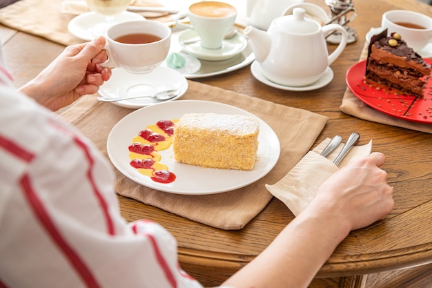 Ragazza che beve il tè con la torta napoleone. una fetta di torta napoleone su un piatto bianco su un tavolo servito. Sullo sfondo c'è un bollitore, tè, succo di frutta.