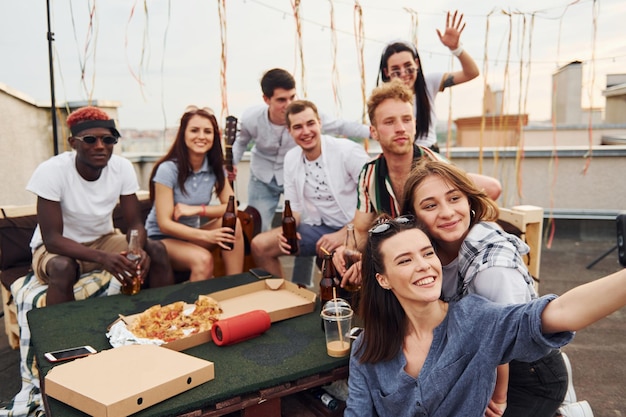 Ragazza che fa selfie con una deliziosa pizza Un gruppo di giovani in abiti casual fanno una festa sul tetto insieme durante il giorno