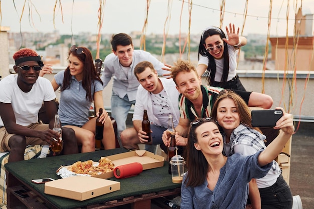 Ragazza che fa selfie con una deliziosa pizza Un gruppo di giovani in abiti casual fanno una festa sul tetto insieme durante il giorno