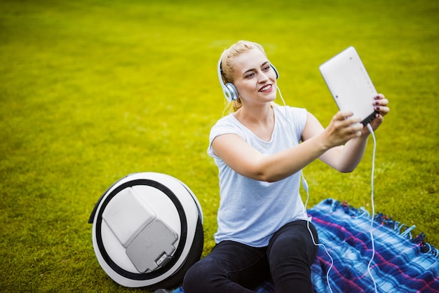 Ragazza che fa selfie sulla compressa che si siede sull'erba in parco.