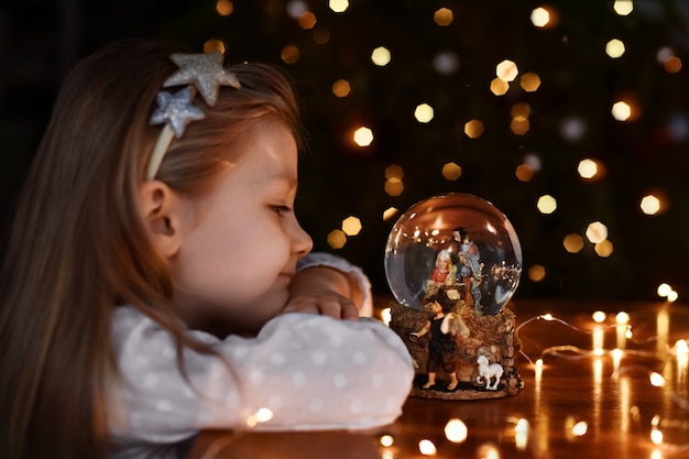 Ragazza che guarda una palla di vetro con una scena della nascita di Gesù Cristo vicino all'albero di natale