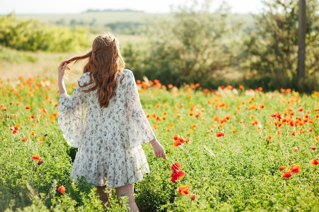 Ragazza che indossa abito bianco e cappello di paglia cammina tra i fiori di papavero rosso al tramonto. La giovane donna gode del buon tempo e si diverte sul campo pieno di fiori rossi. Concetto di primavera e natura