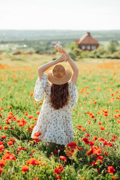 Ragazza che indossa abito bianco e cappello di paglia cammina tra i fiori di papavero rosso al tramonto. La giovane donna gode del buon tempo e si diverte sul campo pieno di fiori rossi. Concetto di primavera e natura