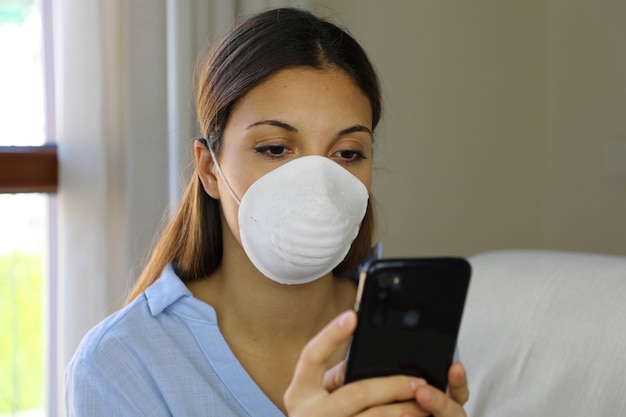 Ragazza che indossa una maschera facciale utilizzando il telefono cellulare che legge le informazioni sulla malattia da Coronavirus 2019 a casa.