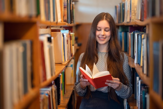 Ragazza che legge un libro con copertina rossa in biblioteca Allegra studentessa bruna che si diverte a leggere un testo affascinante e ad ottenere il piacere dell'istruzione