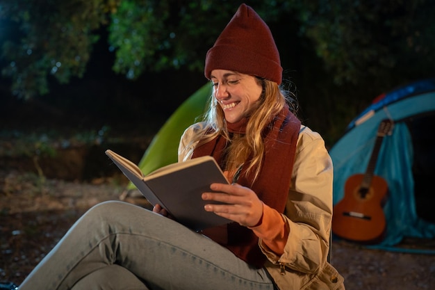 Ragazza che legge un libro di notte in campeggio scena notturna accanto al fuoco del campeggio rilassandosi nella natura tranquilla