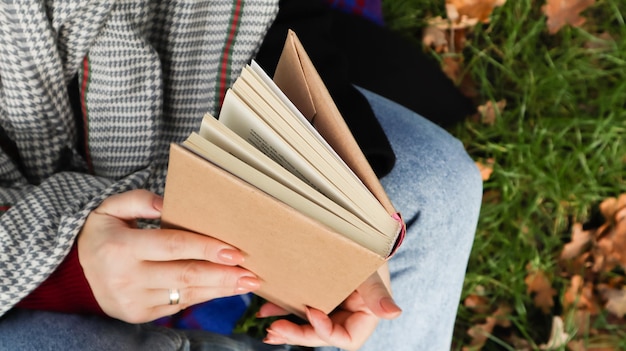 Ragazza che legge un libro nel parco autunnale. Le mani femminili aprono le pagine di un libro di carta all'aperto in una calda giornata di sole. Lo studente si sta preparando per l'esame. Tempo libero letterario in natura.