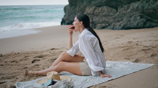 Ragazza che mangia mela a spiaggia picnic seduta a una coperta donna godere di frutta sulla riva del mare