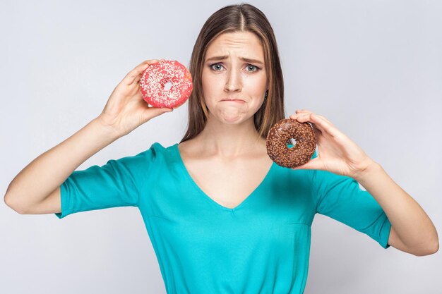 Ragazza che mostra ciambelle rosa e cioccolato con faccia infelice su sfondo grigio