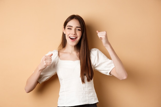 Ragazza che ottiene motivazione, fa pompe a pugno e canta sì, sorride felice, celebra la vittoria o esulta, in piedi su sfondo beige.