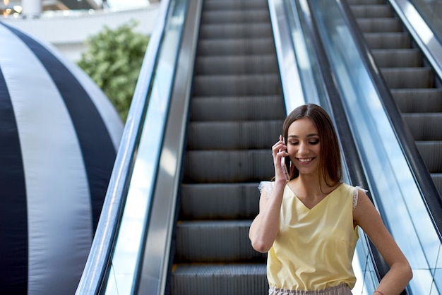 Ragazza che parla al telefono nel centro commerciale che sale la scala mobile