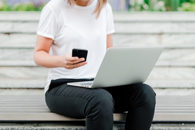 Ragazza che per mezzo del telefono e del computer portatile per lavoro a distanza all'aperto