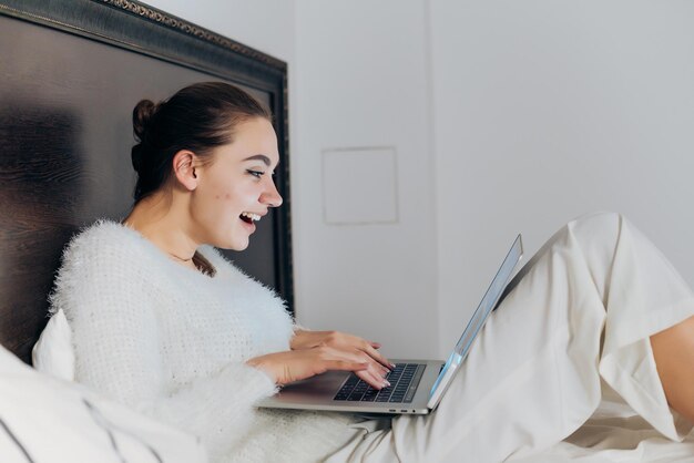 Ragazza che ride felice in vestiti bianchi che si siede a letto e che osserva serie divertenti sul suo computer portatile