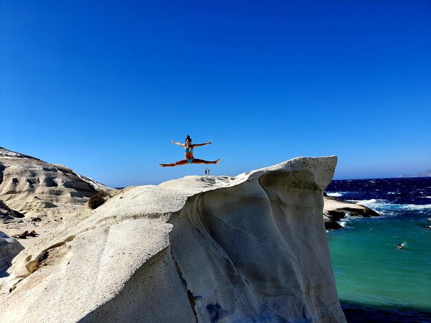 Ragazza che salta su una roccia sul mare contro un cielo blu limpido