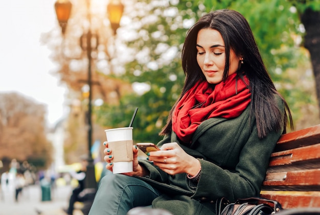 Ragazza che si siede nel parco con caffè e telefono