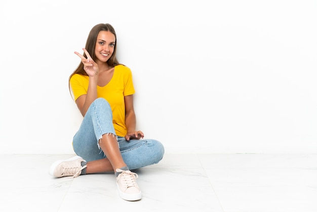 Ragazza che si siede sul pavimento che sorride e che mostra il segno di vittoria