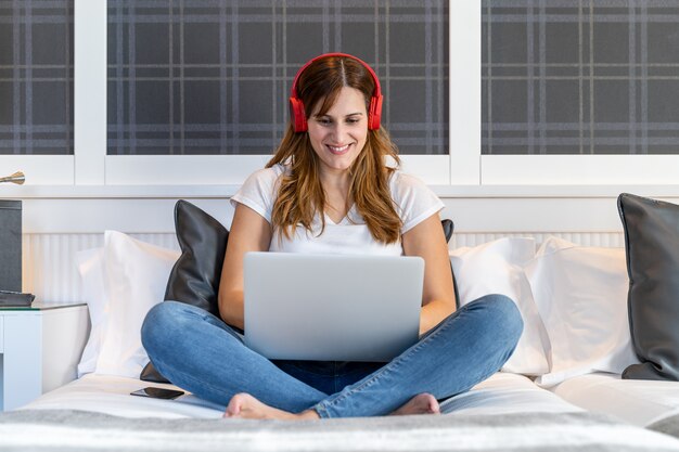 Ragazza che si siede sul suo letto che ascolta la musica e che lavora al suo computer portatile. Concetto di tecnologia e lavoro a casa.