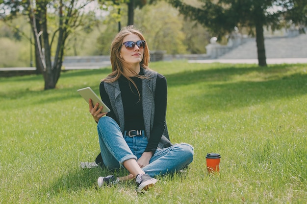 Ragazza che si siede sull'erba con caffè e tablet per strada.