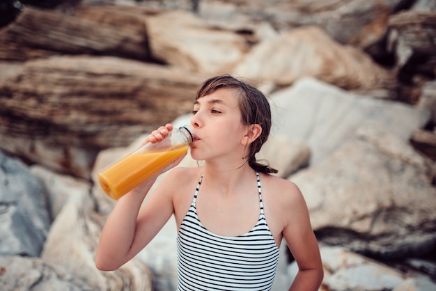 Ragazza che si siede sulla spiaggia e che beve il succo di arancia