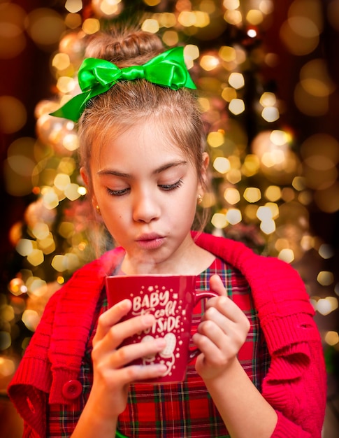 ragazza che soffia in un primo piano della tazza sullo sfondo di un albero di Natale