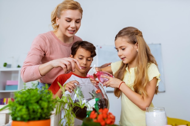 Ragazza che spruzza acqua. Studentessa bionda che spruzza acqua sulle piante grasse in piedi vicino all'allievo e all'insegnante