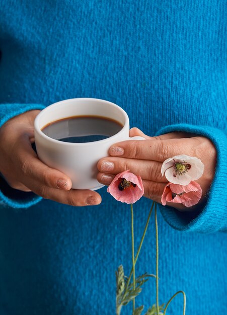 Ragazza che tiene i fiori del papavero e una tazza bianca del caffè caldo di mattina. Sfondo blu.