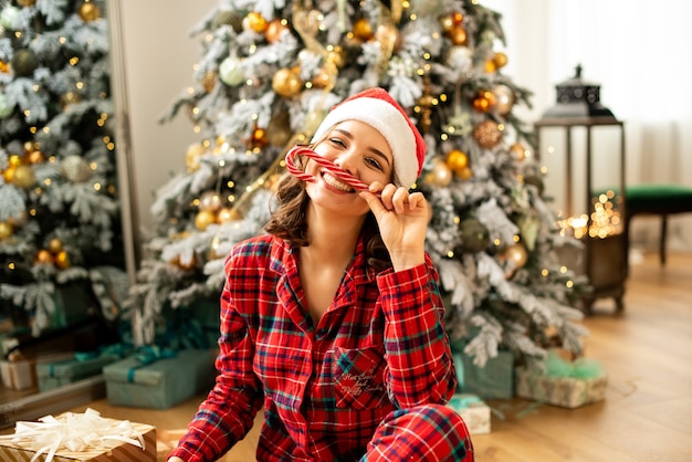 Ragazza che tiene le corna da caramelle di Capodanno e festeggia il Natale. Sullo sfondo albero di Natale decorato con regali.