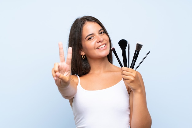 Ragazza che tiene molta spazzola di trucco che sorride e che mostra il segno di vittoria