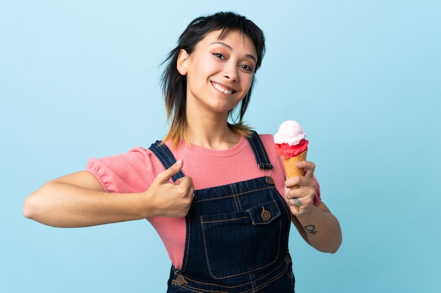 Ragazza che tiene un gelato della cornetta sopra la parete blu con i pollici su perché è successo qualcosa di buono