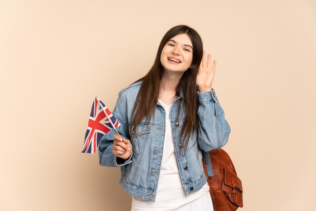 Ragazza che tiene una bandiera del Regno Unito isolata sul beige ascoltando qualcosa mettendo la mano sull'orecchio