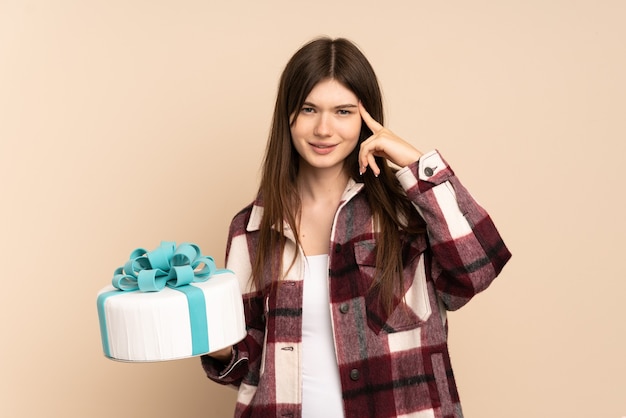 Ragazza che tiene una grande torta isolata sul beige che ha dubbi e pensiero