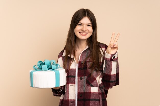 Ragazza che tiene una grande torta isolata sul beige che sorride e che mostra il segno di vittoria